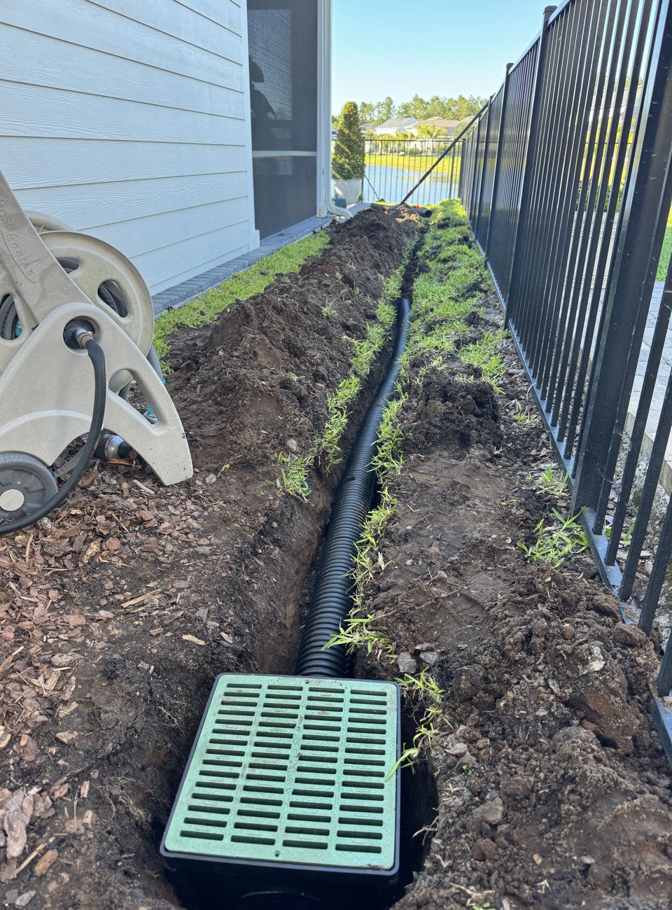Catch basin with a green grate set into a trench beside a home, with corrugated drain pipe running toward a retention pond — yard drainage installation in St. Johns County