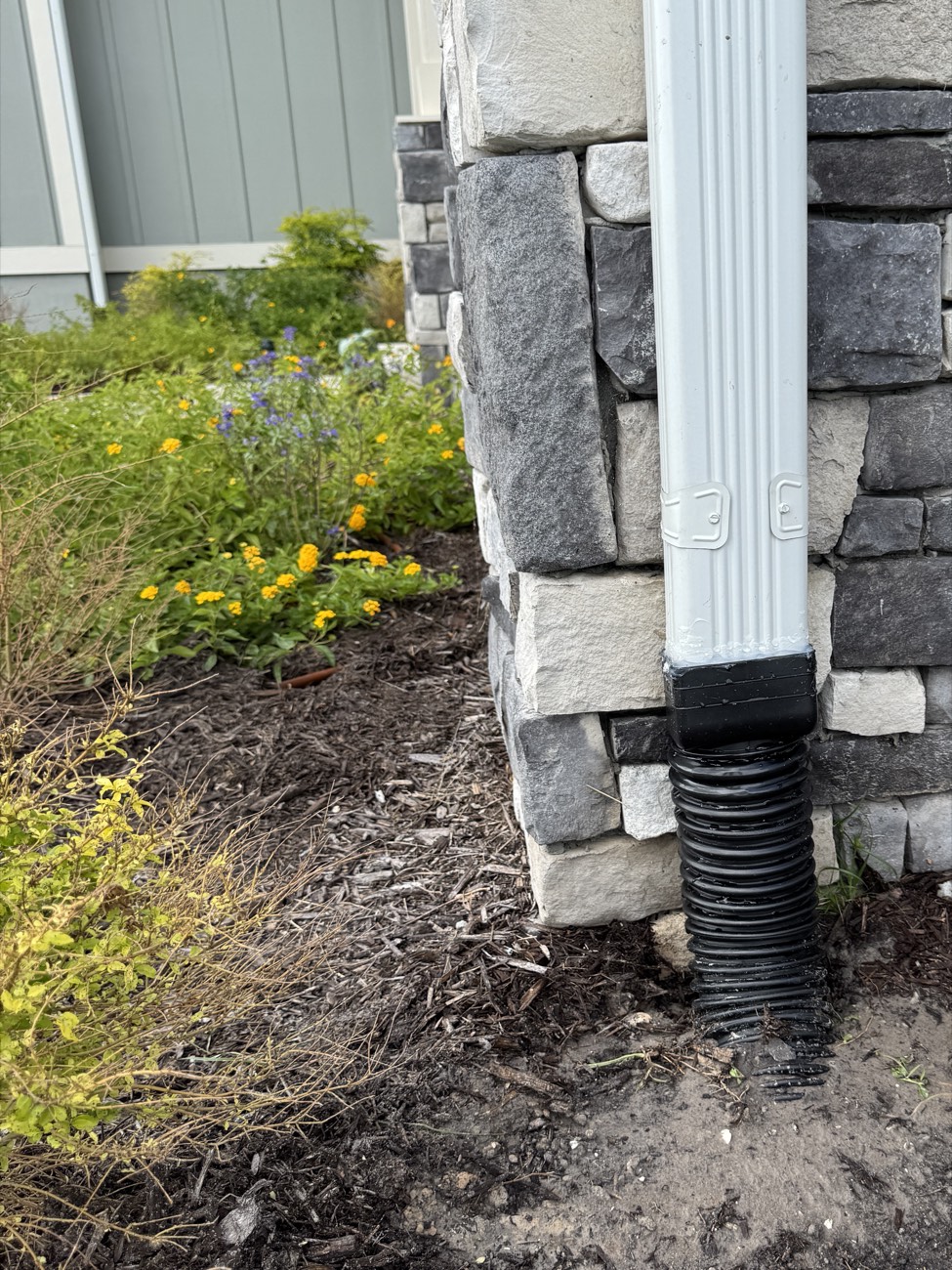 Seamless gutter downspout connected straight into a black corrugated drain line at the base of a stone-veneer wall — gutters and yard drainage tied together on a Northeast Florida home