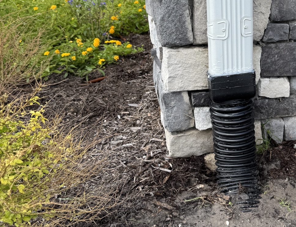 Downspout connected to a buried corrugated gutter drain line beside a stone-clad home in Ponte Vedra Beach